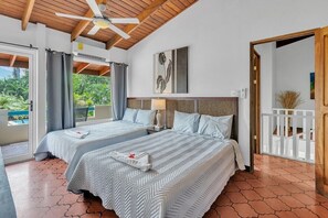 A hotel room with two beds, a ceiling fan, a view of greenery, and a balcony with a white railing.