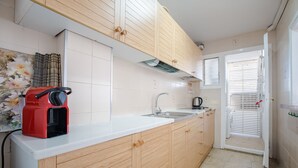 Une cuisine avec des meubles de rangement en bois, un plan de travail blanc, une machine à café rouge et une fenêtre avec des stores.