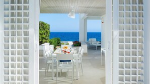 Una terrazza aperta con un tavolo apparecchiato per consumare un pasto, arredi bianchi e vista sul mare.