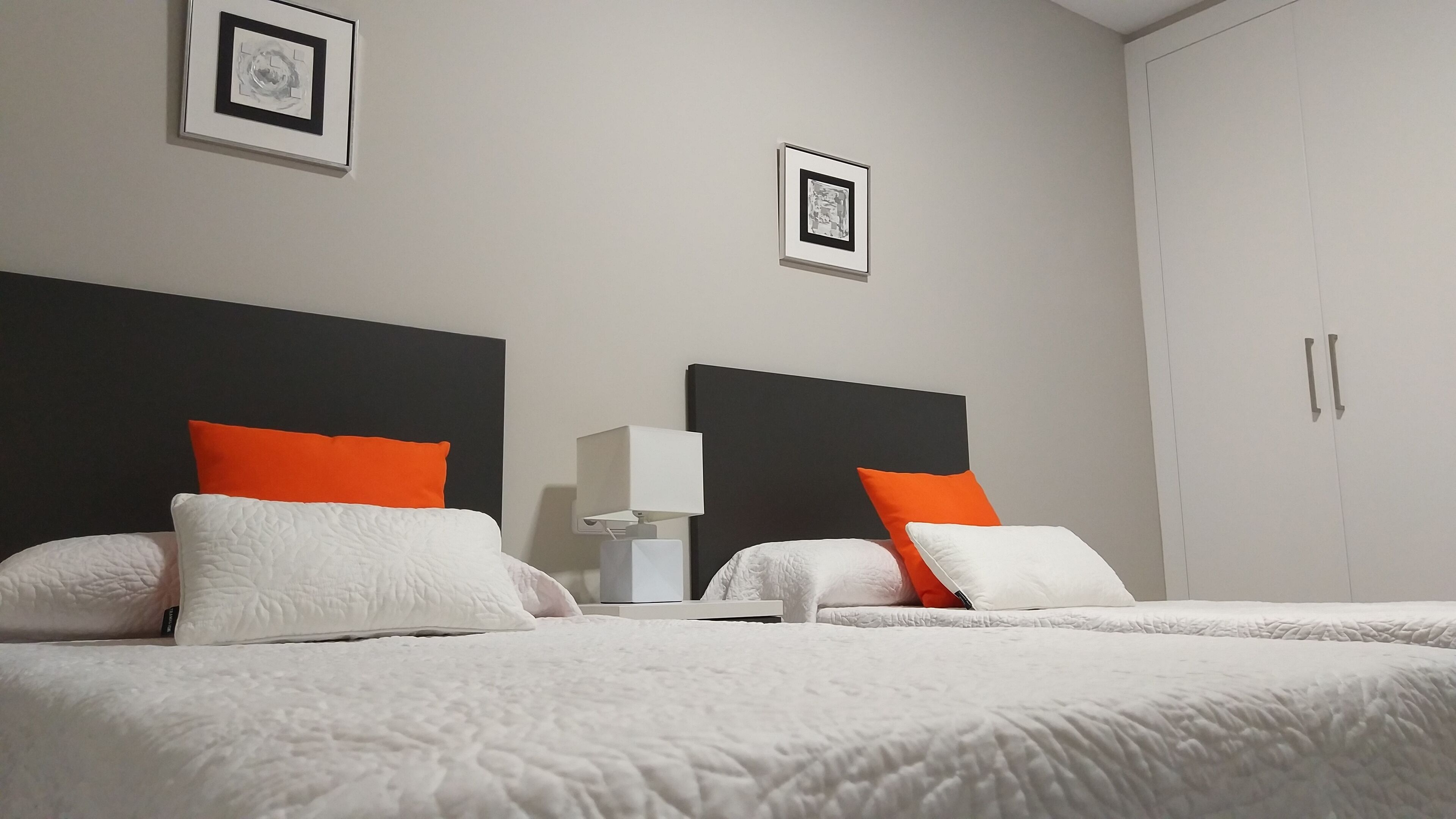 Een hotelkamer met twee bedden, elk voorzien van witte en oranje kussens, een nachtkastje met een lamp, en lijstjes met foto’s aan de muur.