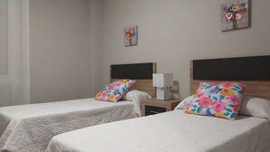 A hotel room with two single beds, each with a floral pillow and a white bedspread. There is a nightstand with a lamp and a small table between the beds. The room has plain walls and a window with sheer curtains. - FULL CENTER REFURBISHED 6 SEATS WIFI. 3 MINUTES FROM LAUREL STREET WALKING (Logroño)