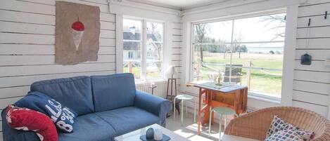A cozy living room with a blue sofa, a wicker chair, a small table, and a view of the outdoors through large windows.