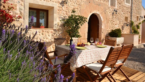 An outdoor dining area with a stone building, wooden chairs, a table with a floral tablecloth, and a potted plant.