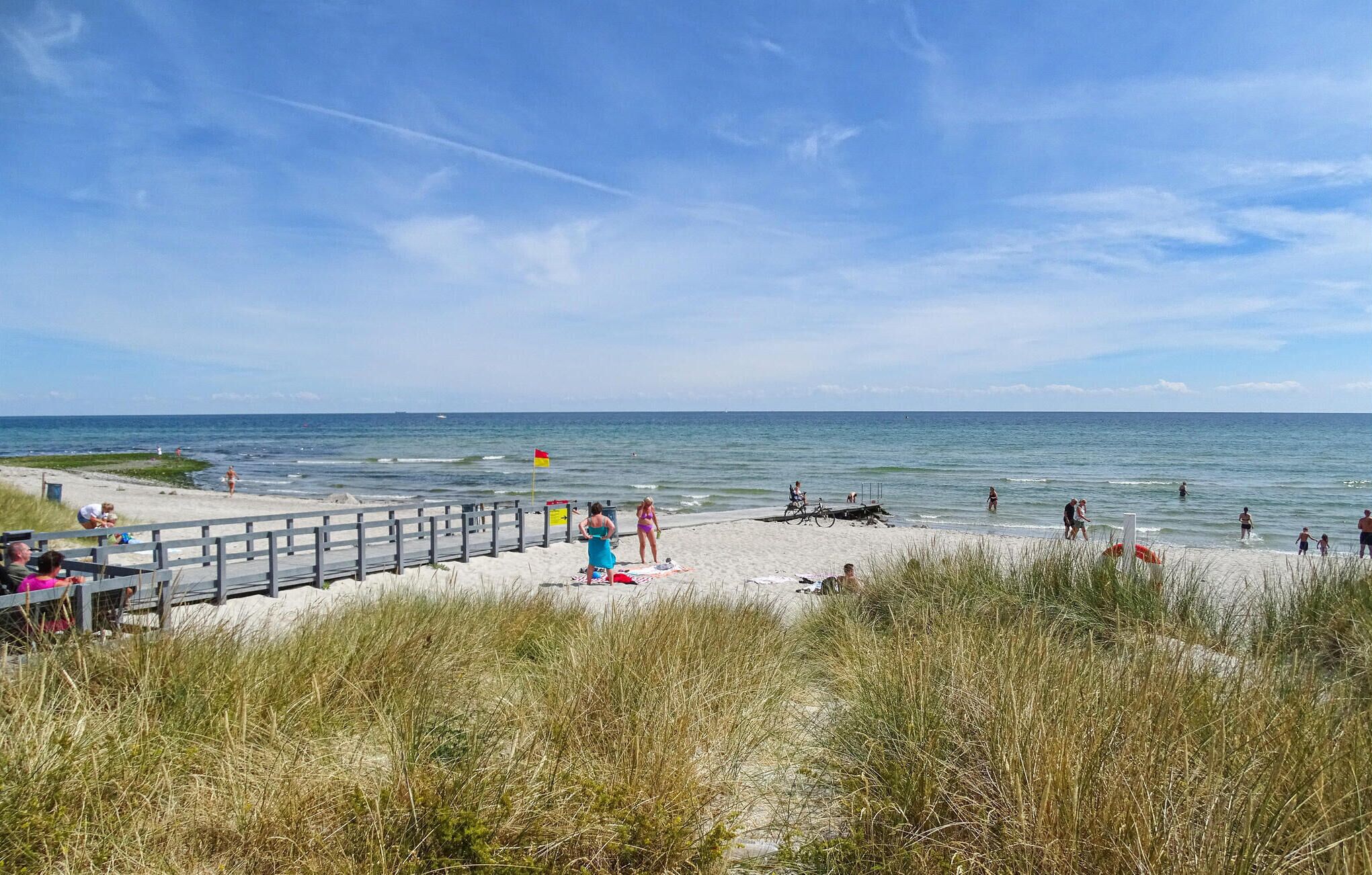 En strand med en träpromenad, människor på sanden och havet i bakgrunden.