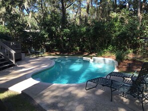 Une piscine à bord incurvé, entourée d’une terrasse et de chaises, située dans un espace boisé.