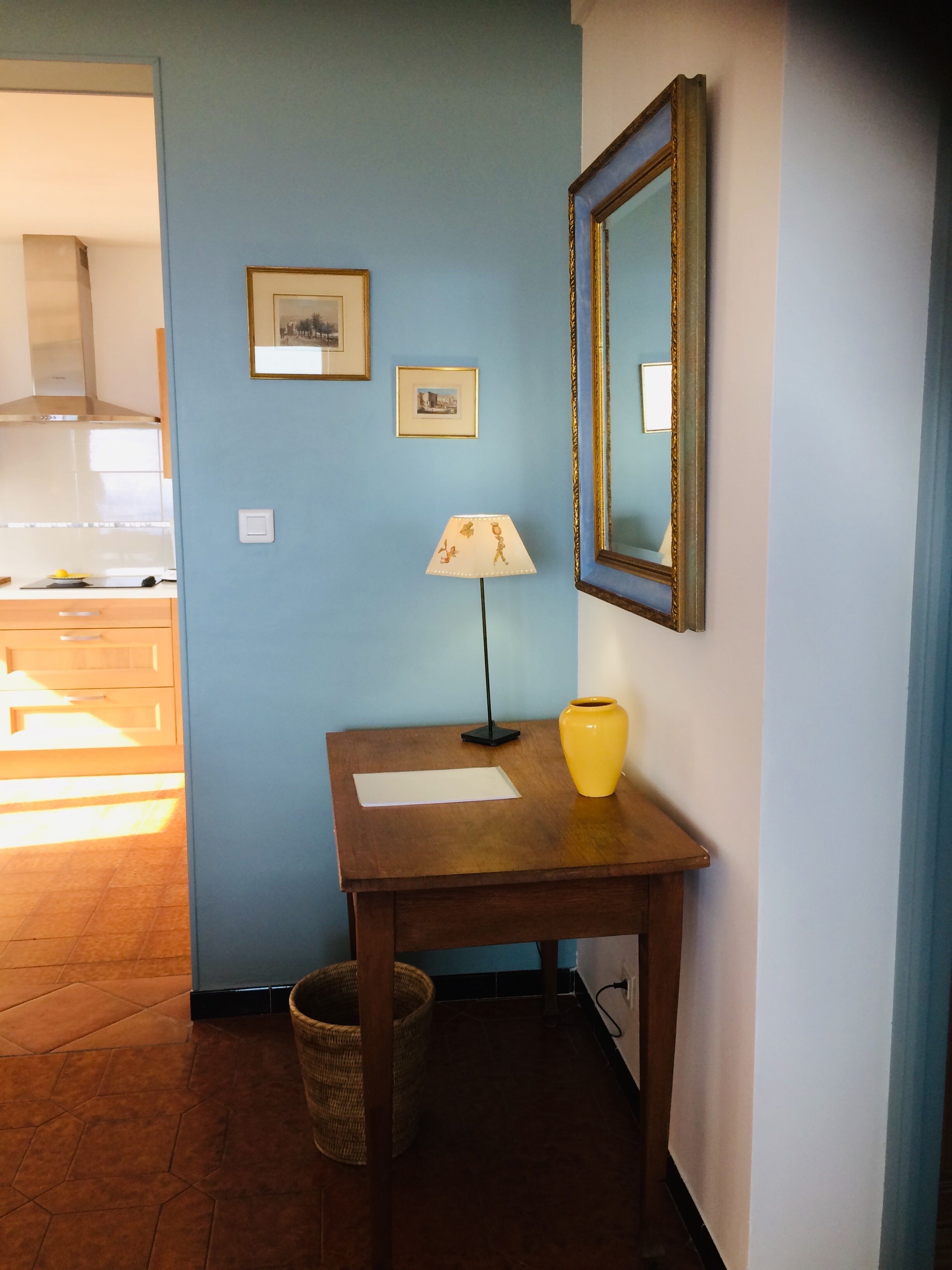 Quarto com parede azul, mesa de madeira com abajur e vaso, espelho e duas imagens emolduradas.