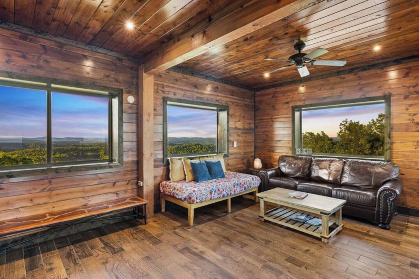 A log cabin interior with wooden walls, large windows, a sofa, a coffee table, and a ceiling fan.