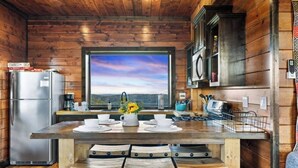 Une cuisine rustique dans un chalet, avec un intérieur en bois, une grande fenêtre offrant une vue panoramique, un réfrigérateur en acier inoxydable et une table à manger en bois avec des chaises.