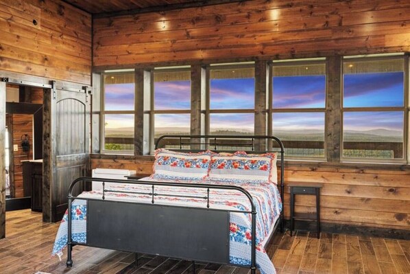 Un dormitorio de cabaña de madera con cama de metal, ventanas amplias y vistas al campo.