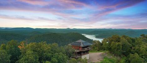A lodge with a single-story structure, surrounded by dense forest, overlooking a lake with mountains in the distance.
