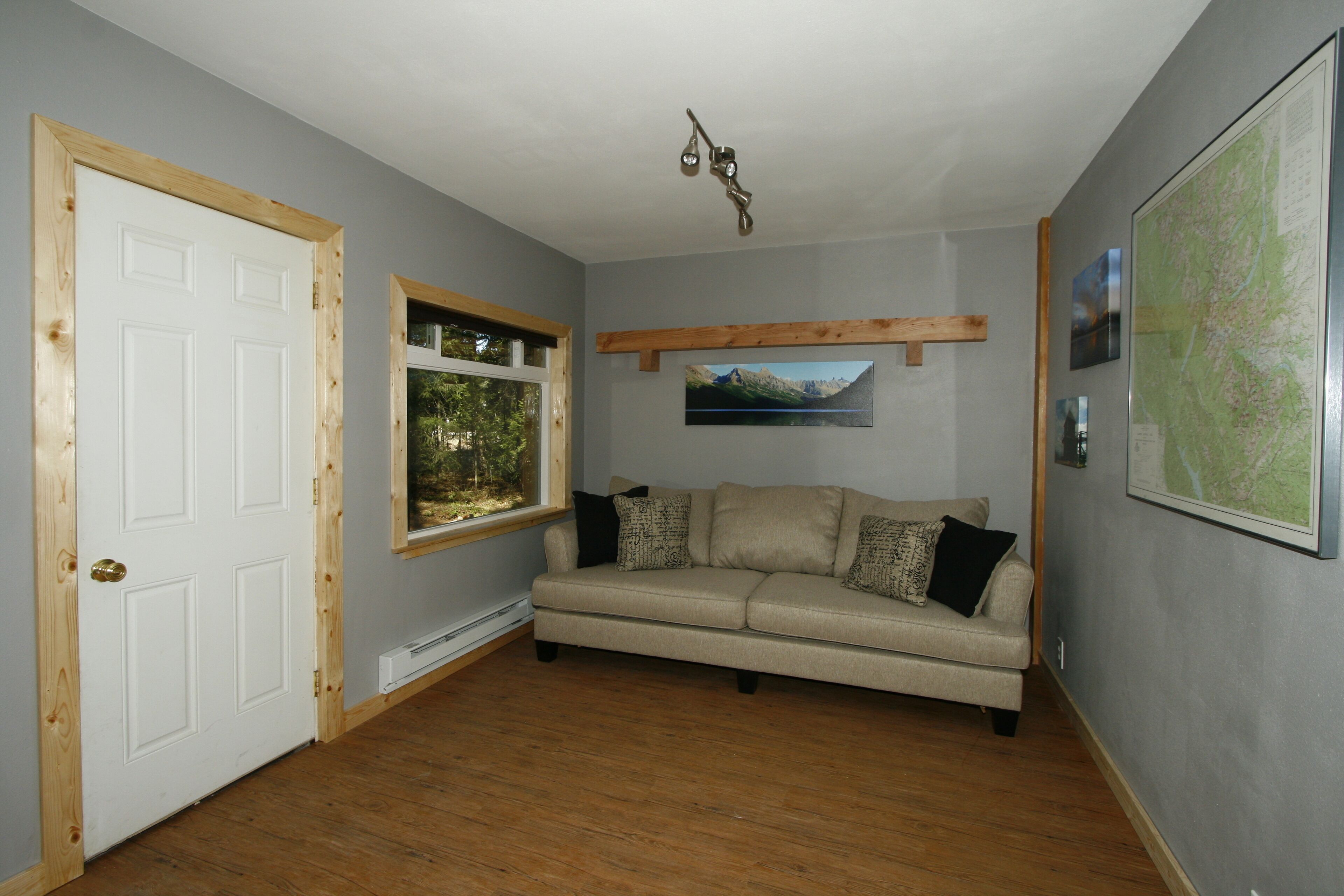 A room with a wooden door, a window with a view of greenery, a beige sofa with cushions, a mounted picture, and a map on the wall.