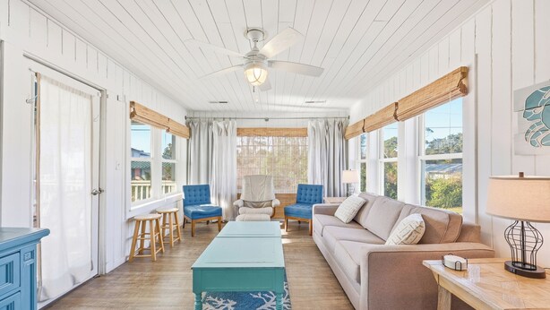 Ein geräumiges Wohnzimmer mit Deckenventilator, einer Couch, einem Couchtisch und Blick ins Freie durch große Fenster.