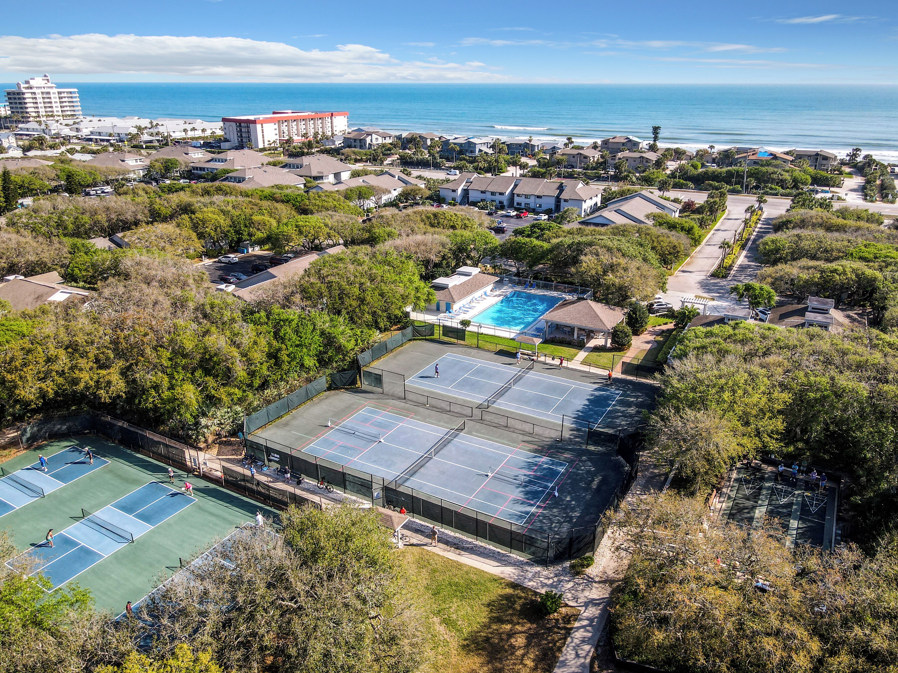 An aerial view of a residential area with a large swimming pool, tennis courts, and a beachfront.