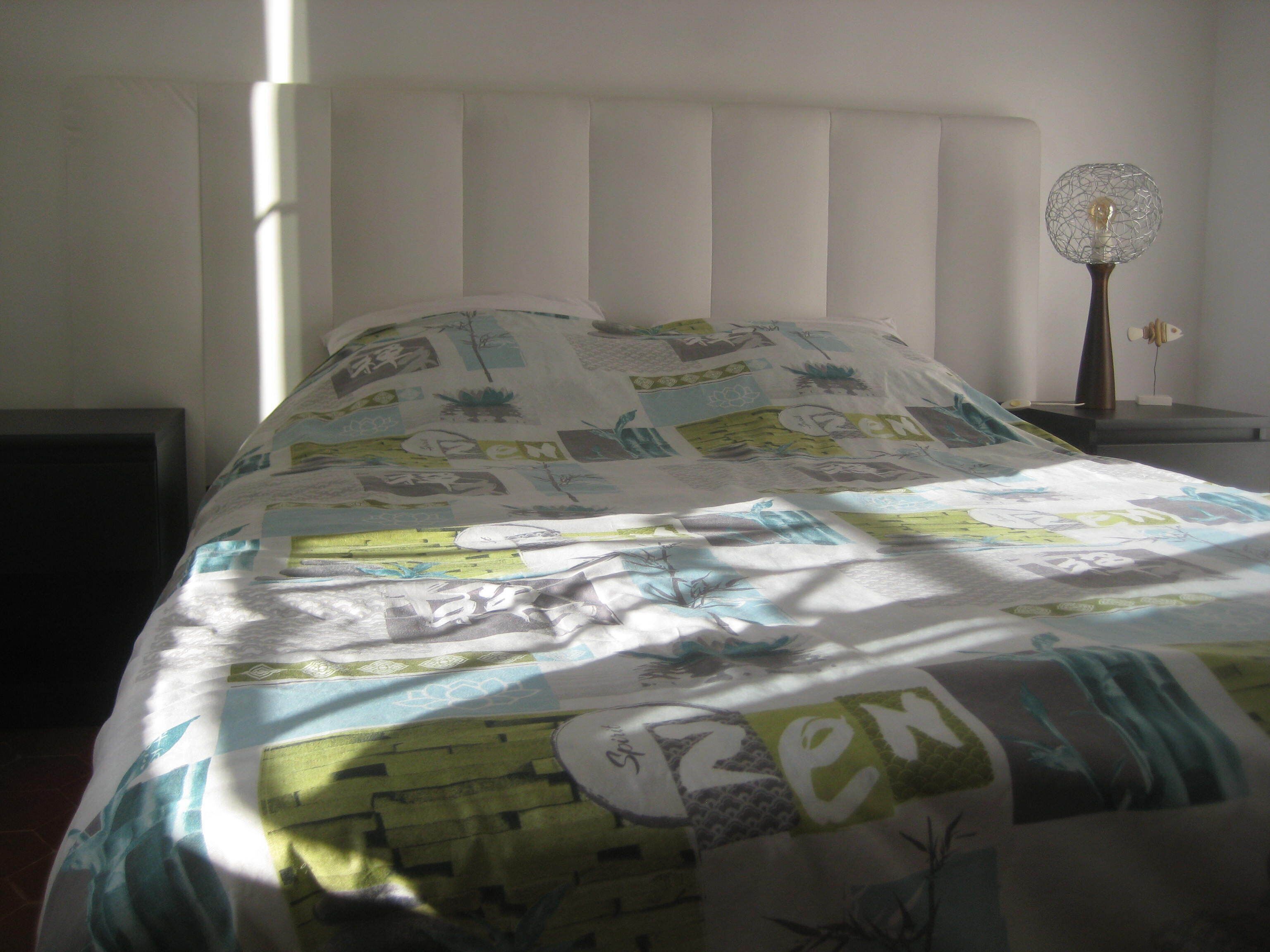 A bed with a quilt featuring various patterns and text, a bedside lamp, and a window allowing natural light.