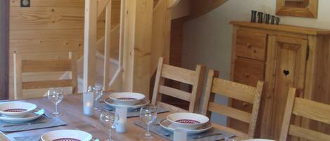 Una mesa de comedor de madera preparada para seis personas, con platos, vasos y cubiertos. Se ve una escalera de madera con barandilla en el fondo.