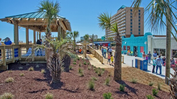 Une promenade en bois avec un kiosque en bois, des palmiers et un bâtiment situé en bord de plage.