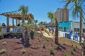 Une promenade en bois avec un kiosque en bois, des palmiers et un bâtiment situé en bord de plage.
