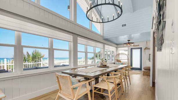 A spacious dining area with a long wooden table and wicker chairs, large windows with a view of the beach, and a ceiling with a circular light fixture.