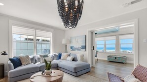 A spacious living room with a large gray sectional sofa, a round wooden coffee table, and a view of the ocean through large windows.