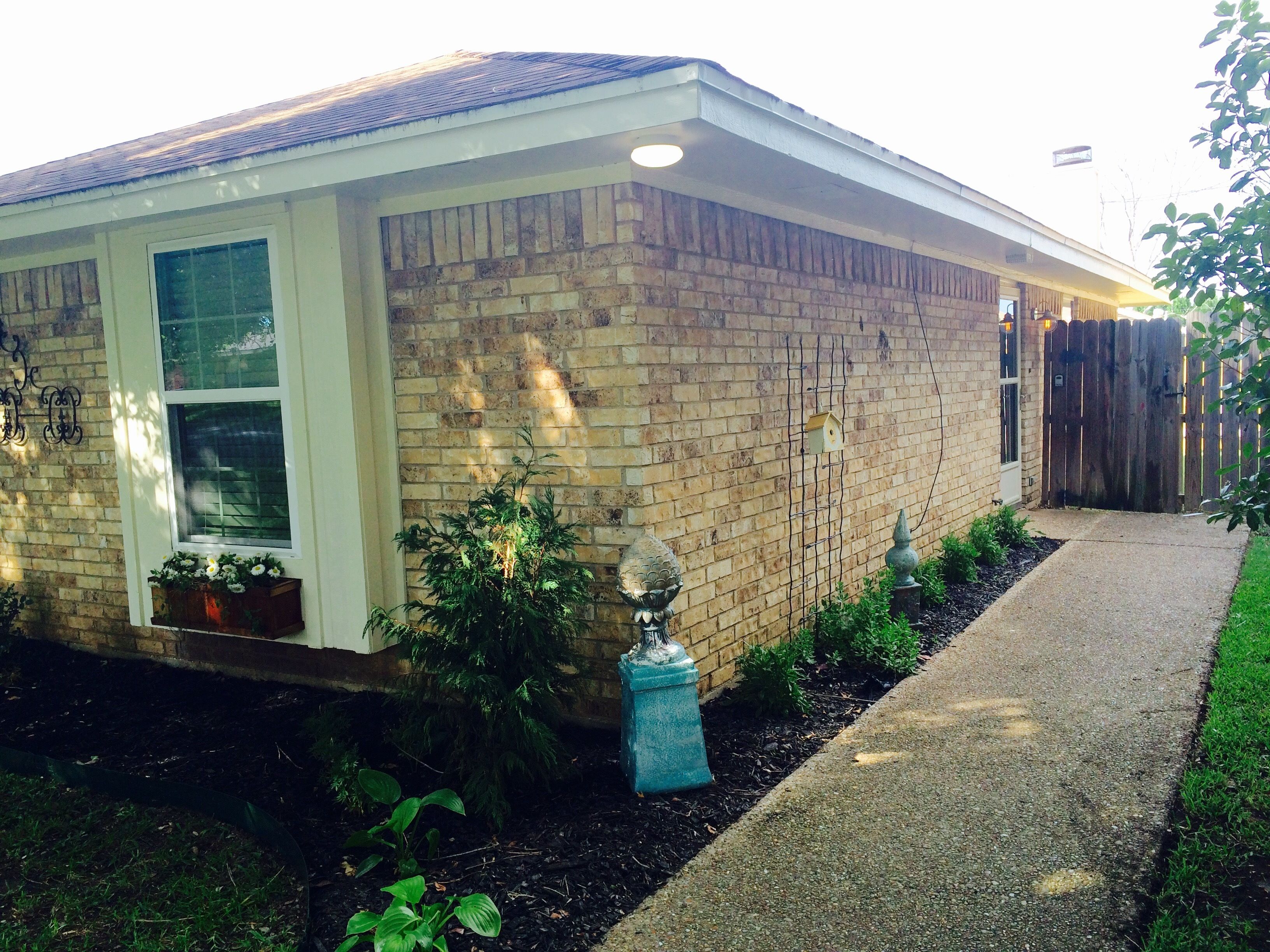 A residential building with a brick exterior, a front door, a window, and a small garden area with plants and a decorative urn.