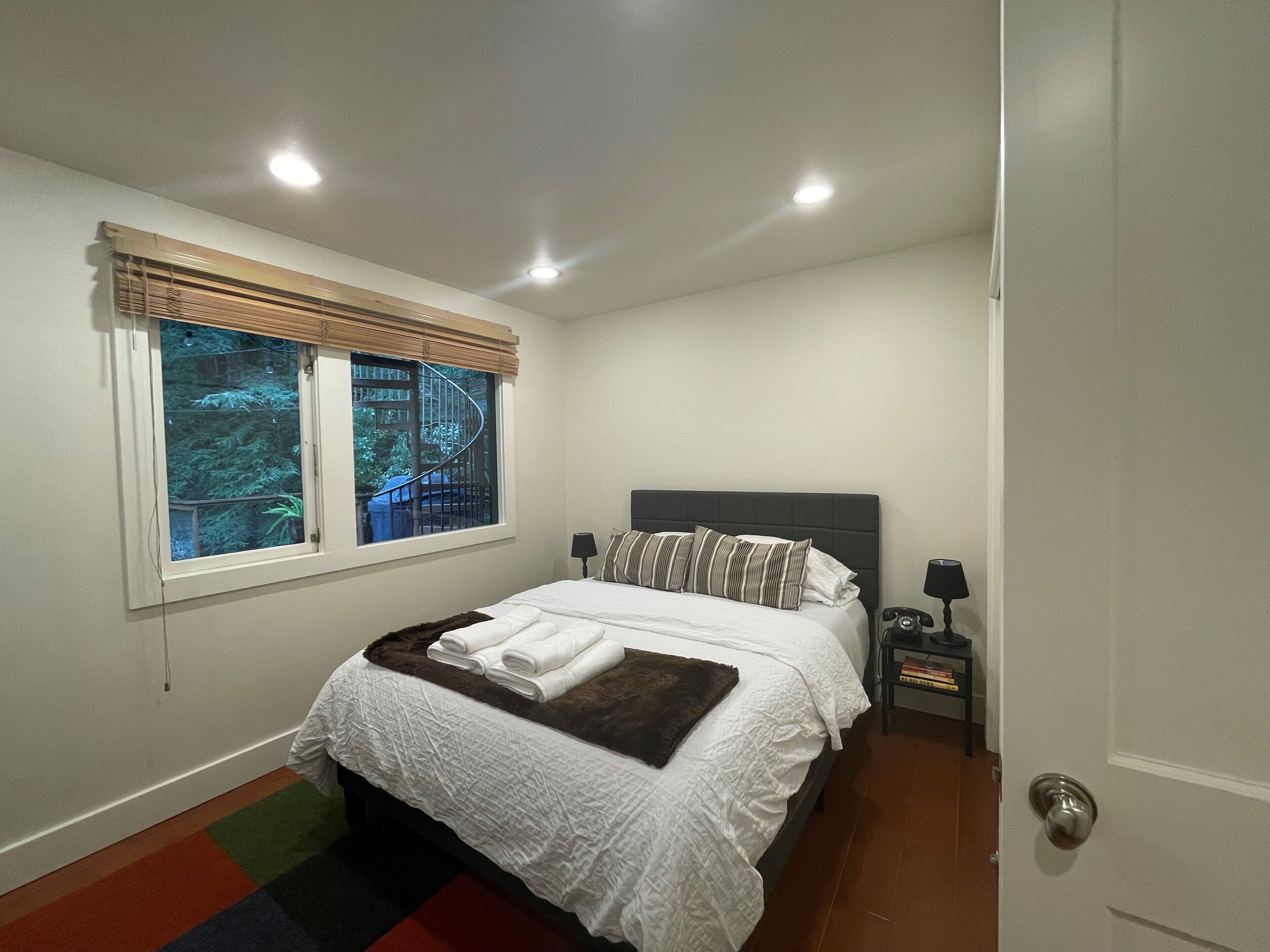 A bedroom with a bed, a window with blinds, a nightstand with a lamp, and a door.
