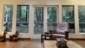 A spacious room with large windows offering a view of a forest. There is a leather armchair and a wooden coffee table with decorative items. - Nestled in the Redwoods, Easy Walk Downtown, Close to SF and Wine Country (Mill Valley)
