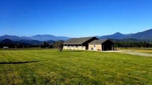 A single-story building with a sloped roof, a small porch, and a door. It is situated in a grassy field with a clear blue sky above and mountains in the background.