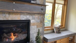 Una chimenea de piedra con un televisor encima, un pequeño árbol de Navidad y un banco con un cojín estampado.