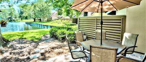 An outdoor patio with a table and chairs, a large umbrella, and a view of a pond and greenery.