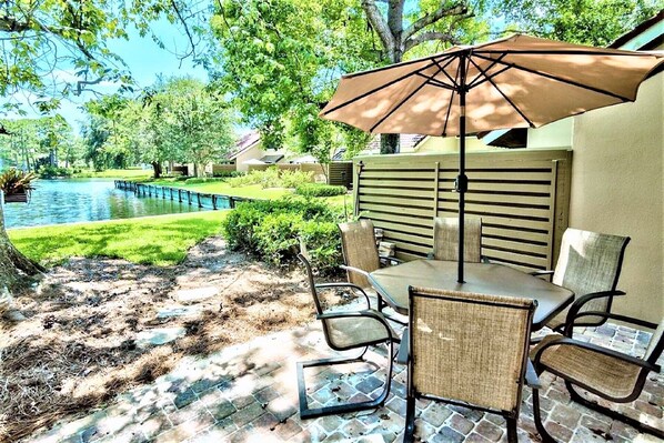 An outdoor patio with a table and chairs, a large umbrella, and a view of a pond and greenery.