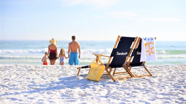 A beach scene with a family walking towards the ocean. There are two beach chairs labeled "Sanderin" and a towel with a logo.