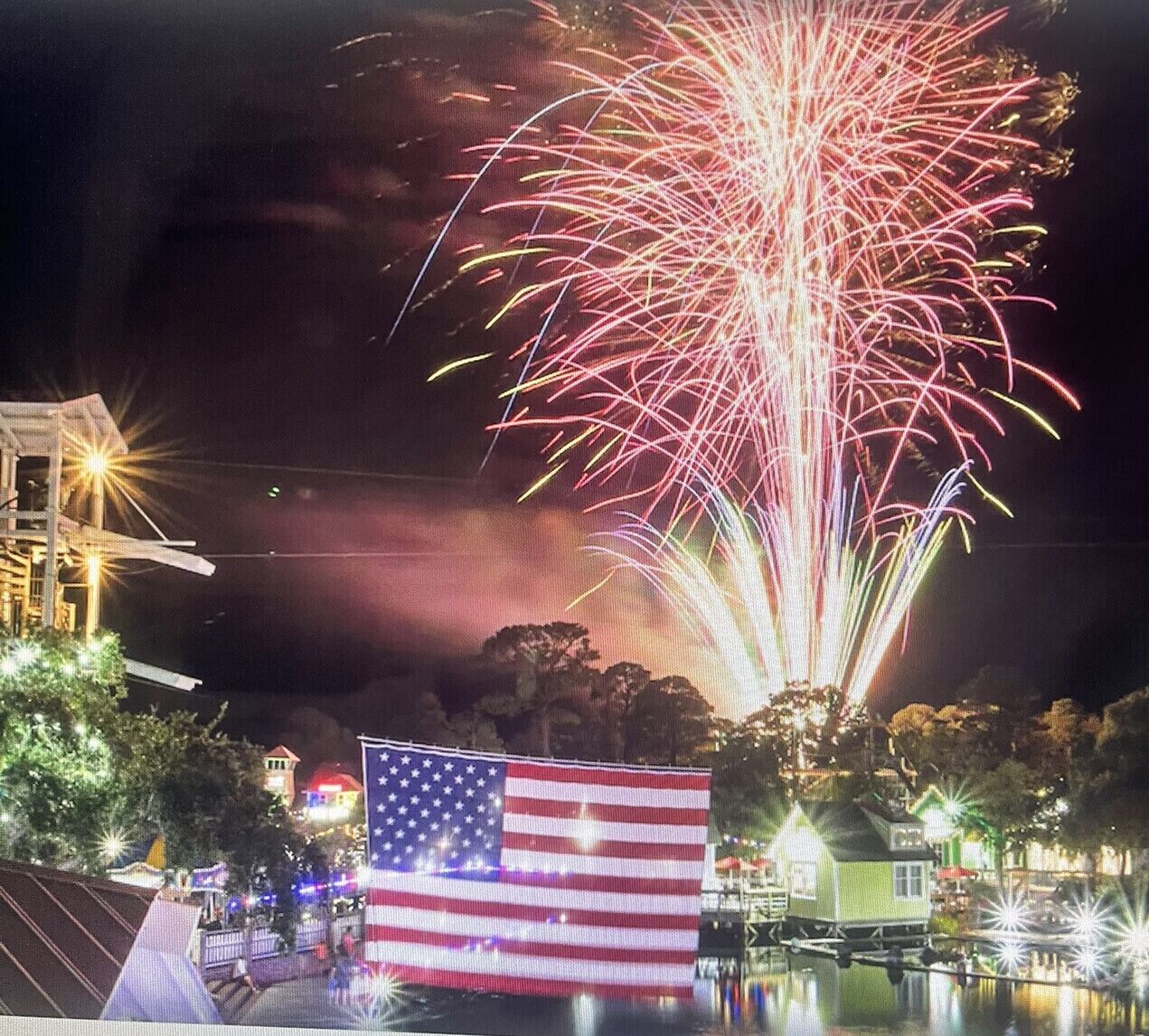 A nighttime scene with fireworks, an American flag, and a small house by a body of water.