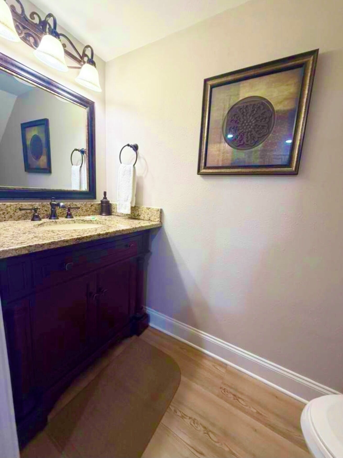 Un baño con tocador, lavabo, espejo, perchero para toallas, cuadro enmarcado y suelo de madera.