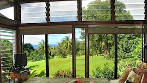 Un dormitorio con un ventanal amplio, una cama con un colchoneta enrollada, una mesita con un televisor y control remoto, y vistas a un jardín exuberante y al océano.