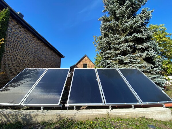 Solar panels installed on a concrete base, a brick building, and a large evergreen tree in the background.