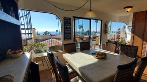 Un coin repas avec des chaises et une table en osier, de grandes fenêtres offrant une vue sur l’océan et un panneau indiquant les principaux sites touristiques de la région.