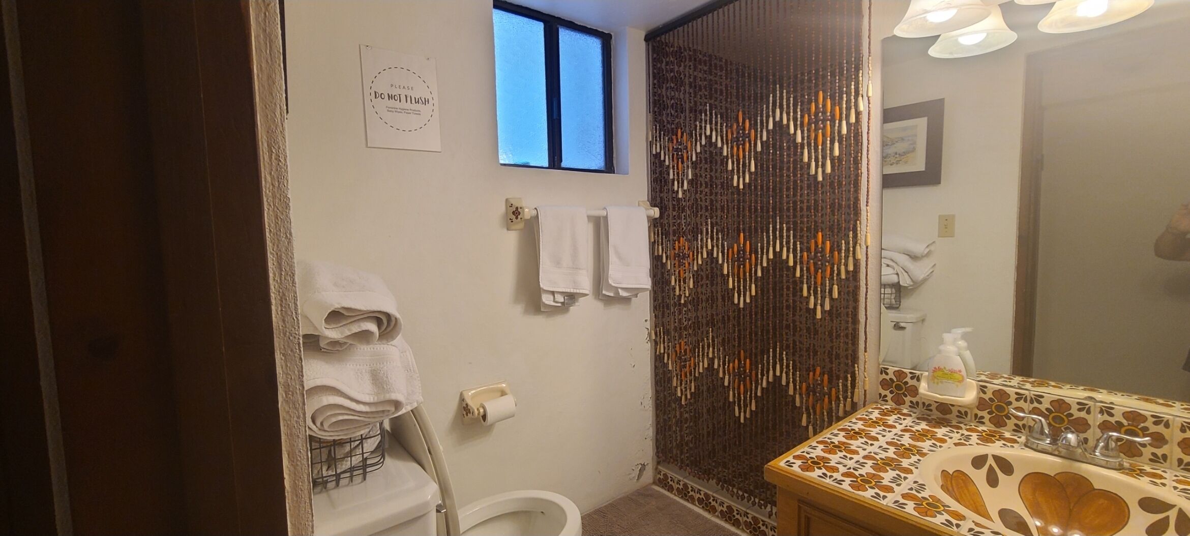 Une salle de bain comprenant des toilettes, un lavabo avec un pare-éclaboussures décoré de motifs floraux, un porte-serviettes avec des serviettes, un panneau indiquant « NE PAS TIRER LA CHAÎNETTE » et un rideau de douche orné de décorations suspendues.