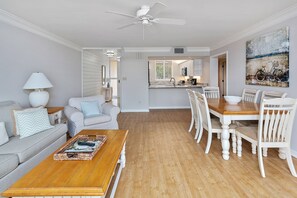 A spacious living room with a wooden coffee table, a grey sofa, and a dining area with a wooden table and white chairs.