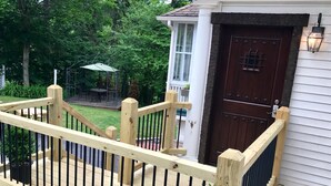 A wooden deck with a railing, a dark brown door, and a small porch area with a table and chairs. - BEST LOCATION! Beautiful Village Home, Walk Everywhere. (Lake Placid)