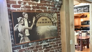 A brick wall with a mural of dancers, wooden beams, and a kitchen area in the background. - BEST LOCATION! Beautiful Village Home, Walk Everywhere. (Lake Placid)