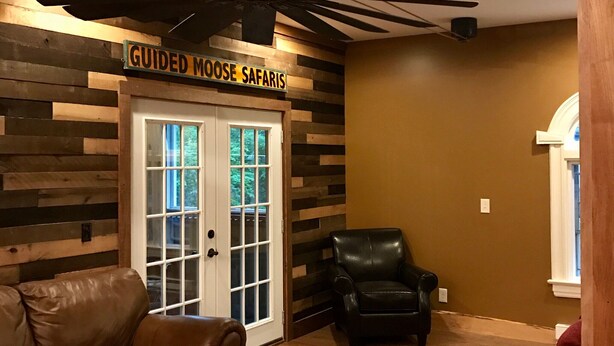 A room with a wooden wall, a ceiling fan, a leather sofa, a black leather chair, and a door labeled "Guided Moose Safaris". - BEST LOCATION! Beautiful Village Home, Walk Everywhere. (Lake Placid)