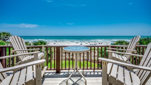 A balcony with two Adirondack chairs and a small table, overlooking a beach with people and palm trees. - Stunning Beach Home! 3 Oceanfront Masters w/ ensuite Baths! Panoramic Views!  (Myrtle Beach)