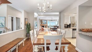 A spacious dining area with a long wooden table and white chairs, a chandelier, and a view of the kitchen and living area. - Stunning Beach Home! 3 Oceanfront Masters w/ ensuite Baths! Panoramic Views!  (Myrtle Beach)
