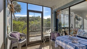 Une pièce baignée de lumière, avec une porte-fenêtre coulissante donnant sur un balcon, un lit recouvert d’une couverture à carreaux bleus et blancs, et une chaise agrémentée d’un coussin violet.