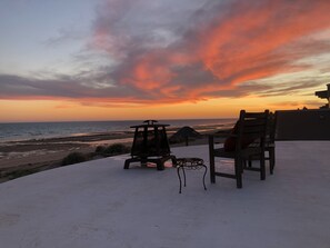 Une terrasse au bord de la plage, avec deux chaises en bois et une petite table, d’où l’on peut admirer l’océan au coucher du soleil.