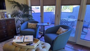 Ein gemütliches Wohnzimmer mit einer Couch, einem Sessel und einem Hocker, einem Couchtisch mit Zeitschriften sowie einem großen Fenster mit Blick auf den Garten.