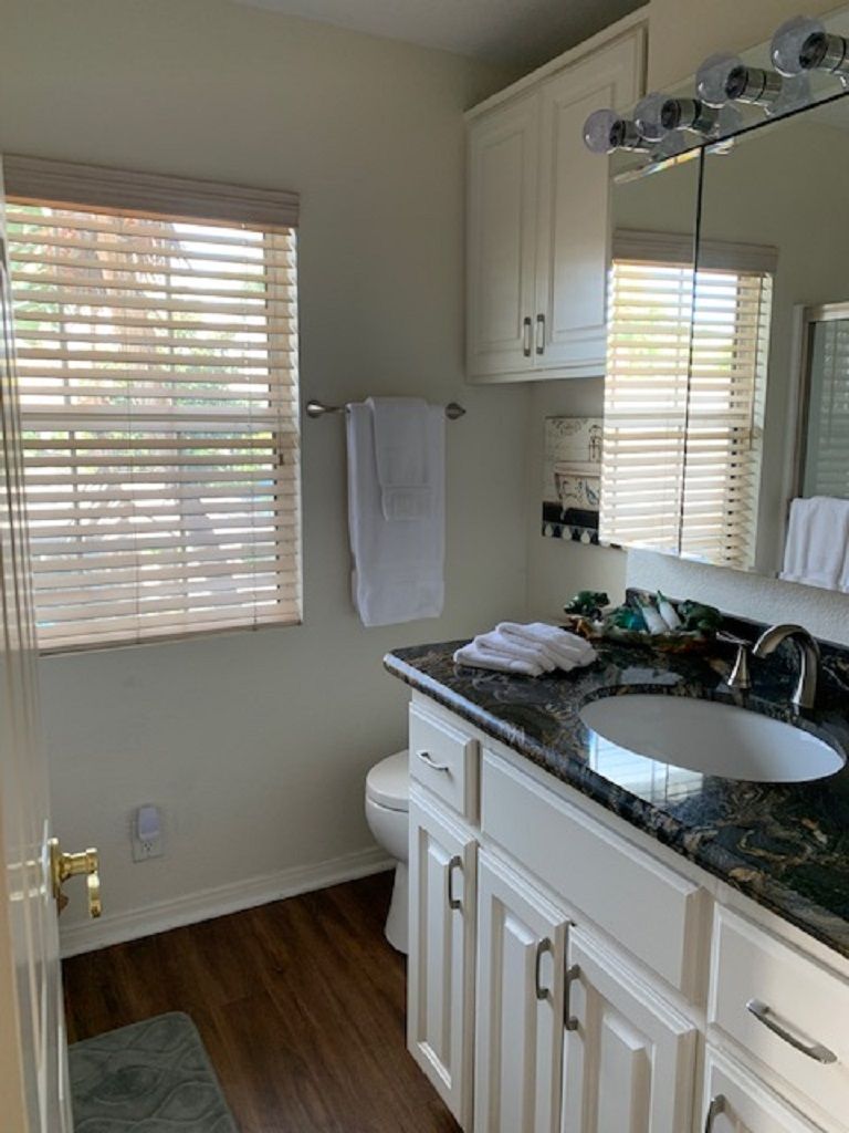 A bathroom with a white sink, vanity, and a large mirror. There is a window with blinds, a towel rack, and a toilet.