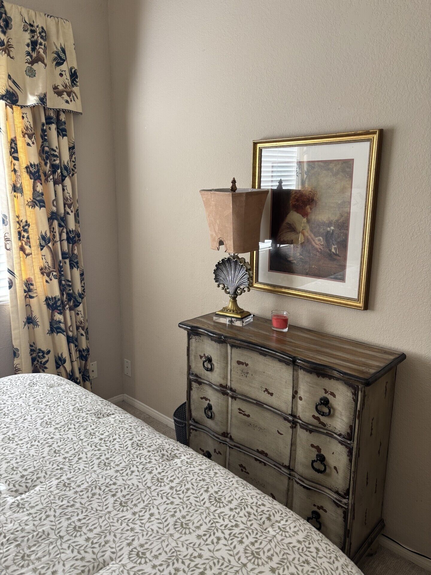 A bedroom with a bed, a dresser with drawers, a lamp, a framed picture on the wall, and curtains with a floral pattern.