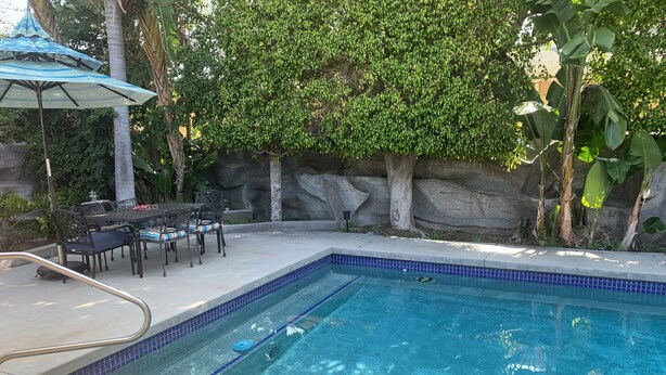 Ein Schwimmbecken mit Fliesenrand, umgeben von einer Terrasse mit Gartenmöbeln und einer Steinwand.