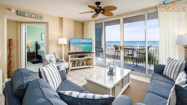 A living room with a sofa set, a coffee table, and a television displaying a beach scene. There is a sign indicating a beach direction and a balcony with a view of the ocean. - Island Princess 214-Beautiful Updated 2Bd Beachfront Condo-Free Beach Service (Fort Walton Beach)